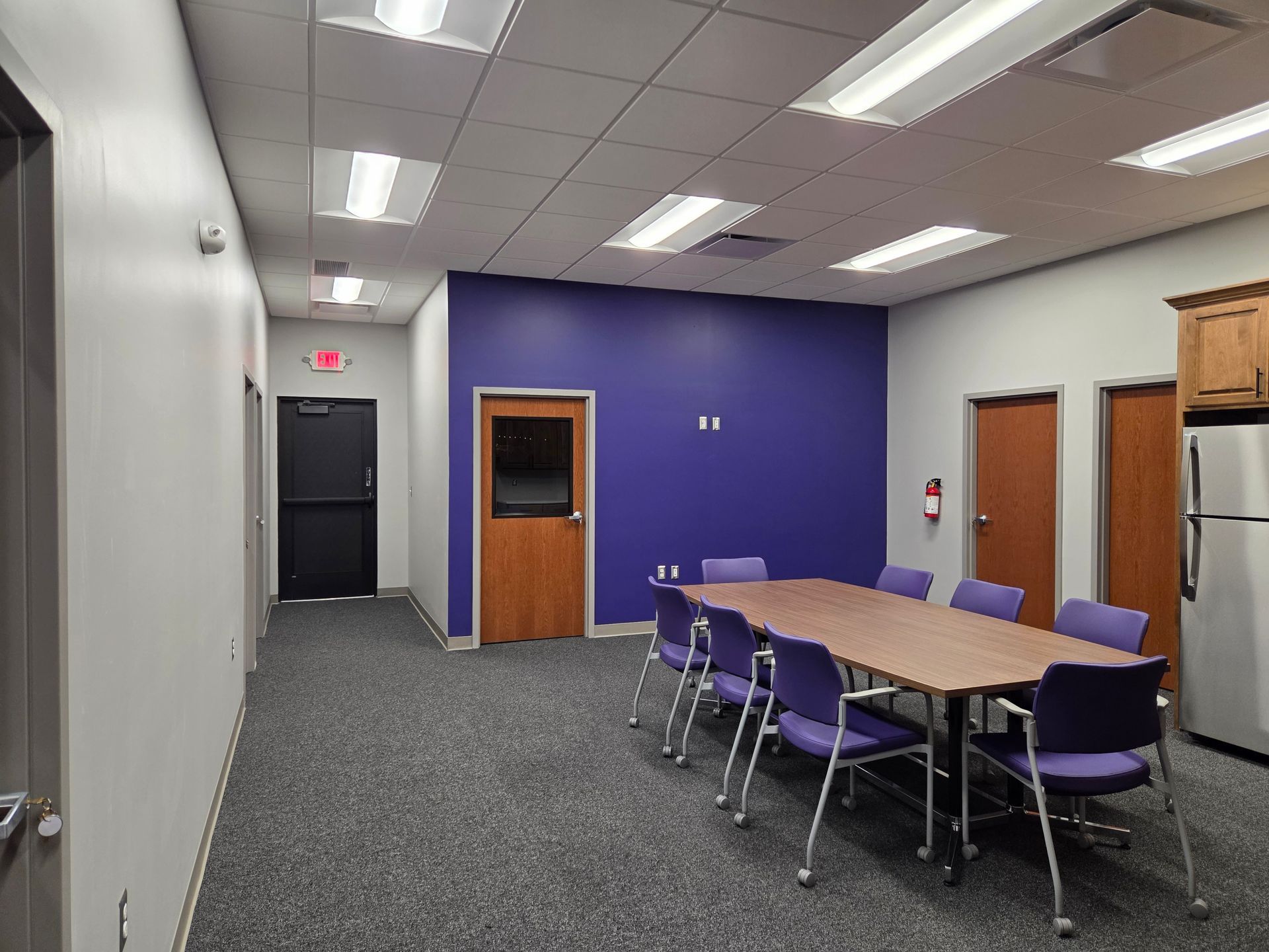 Small meeting room with a purple accent wall, conference table, chairs, and doors along the side.