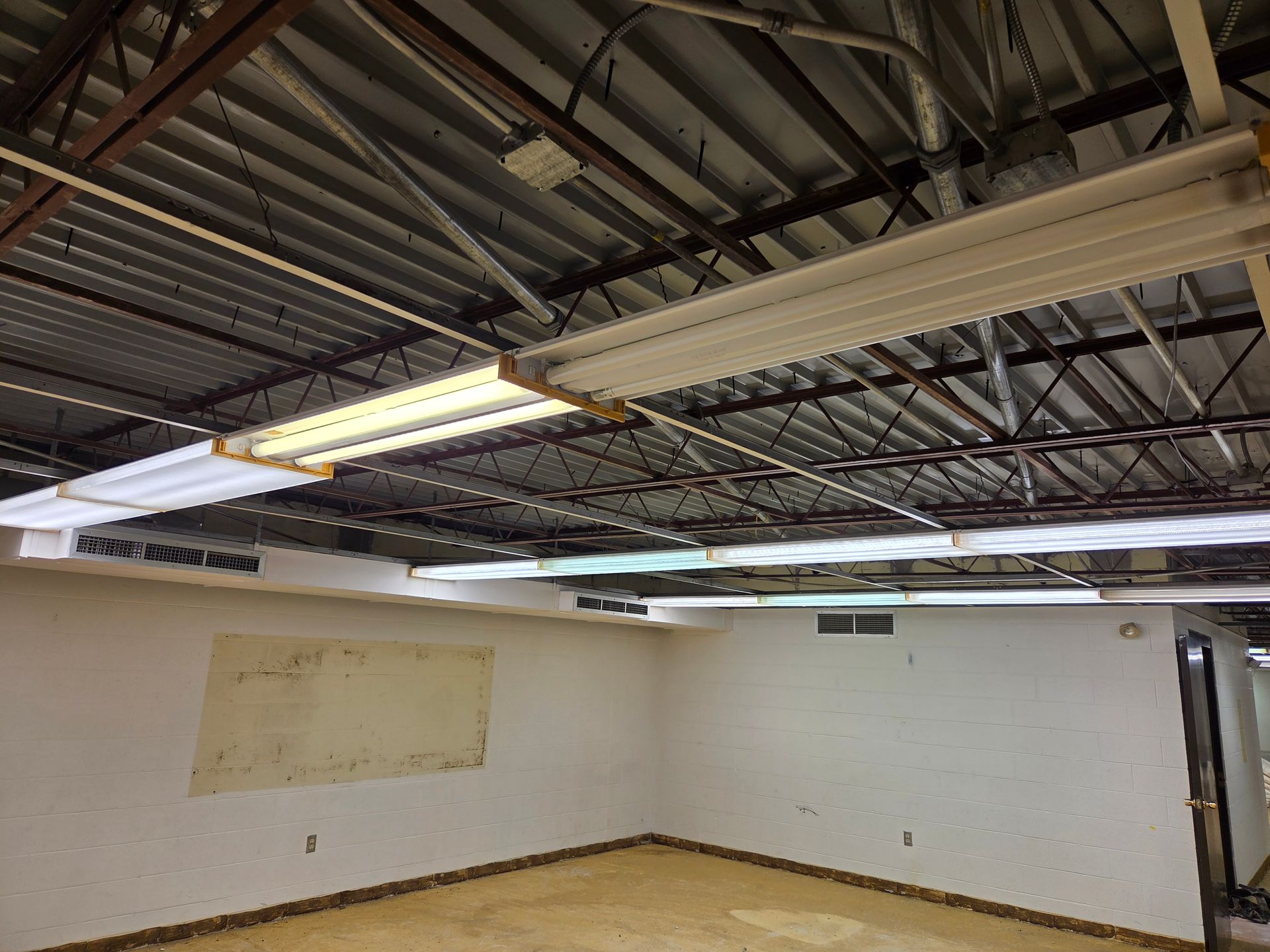 Empty unfinished room with exposed ceiling beams, fluorescent lights, white walls, and a beige floor