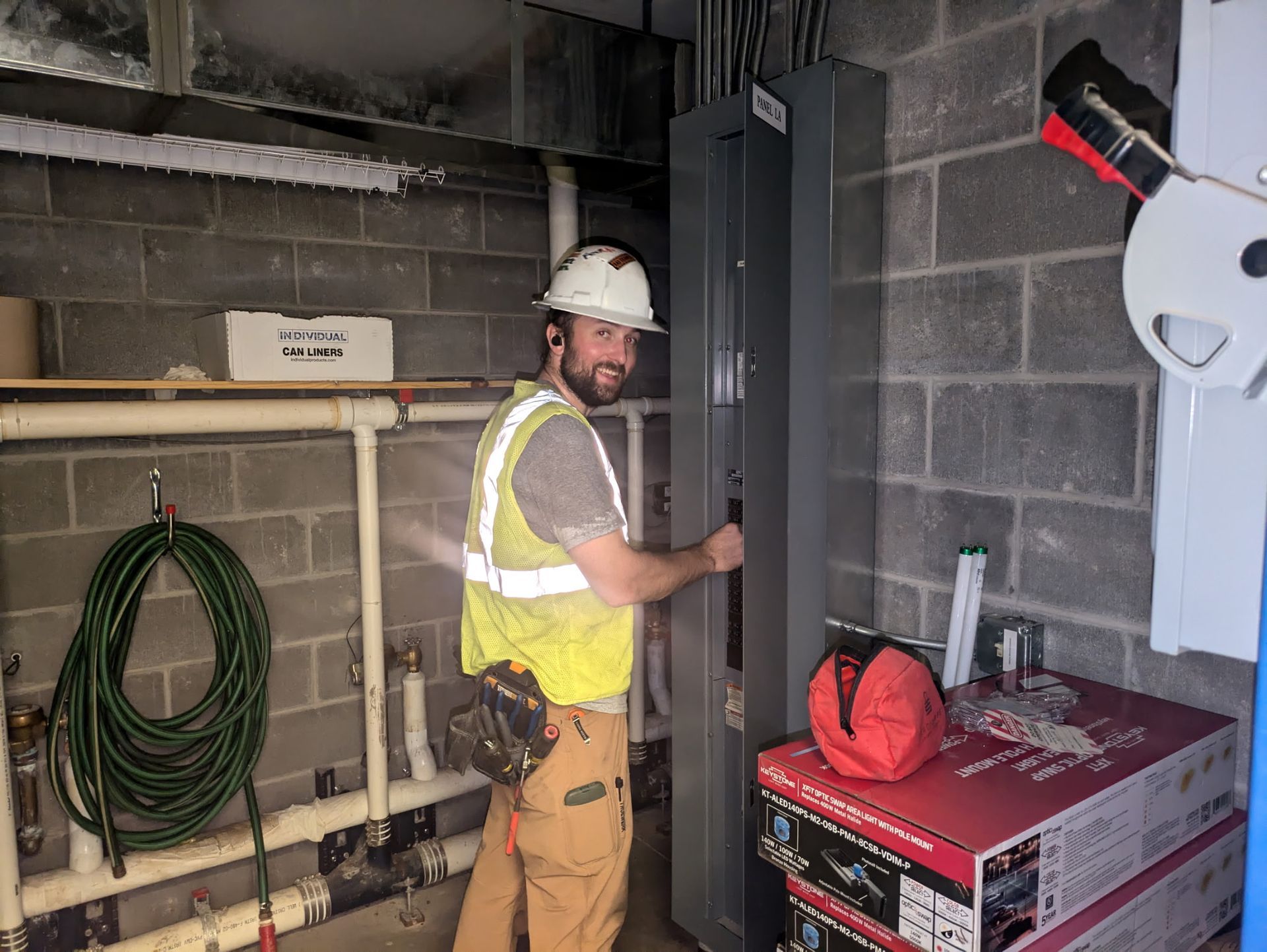 Worker in hard hat and safety vest installing equipment in a utility room with cinder block walls.