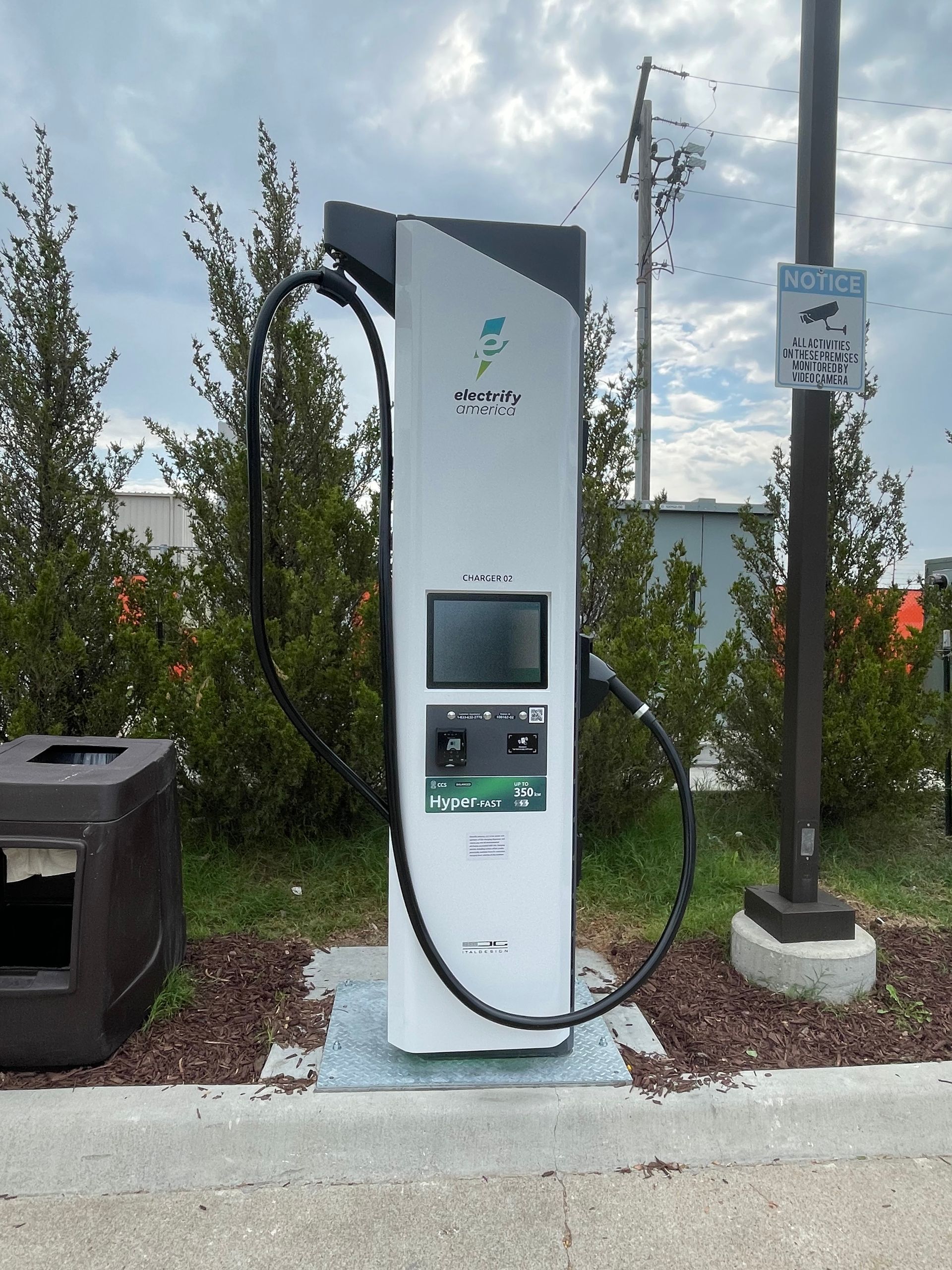 Electric vehicle charging station on a sidewalk beside a street, with a trash bin and utility pole nearby.