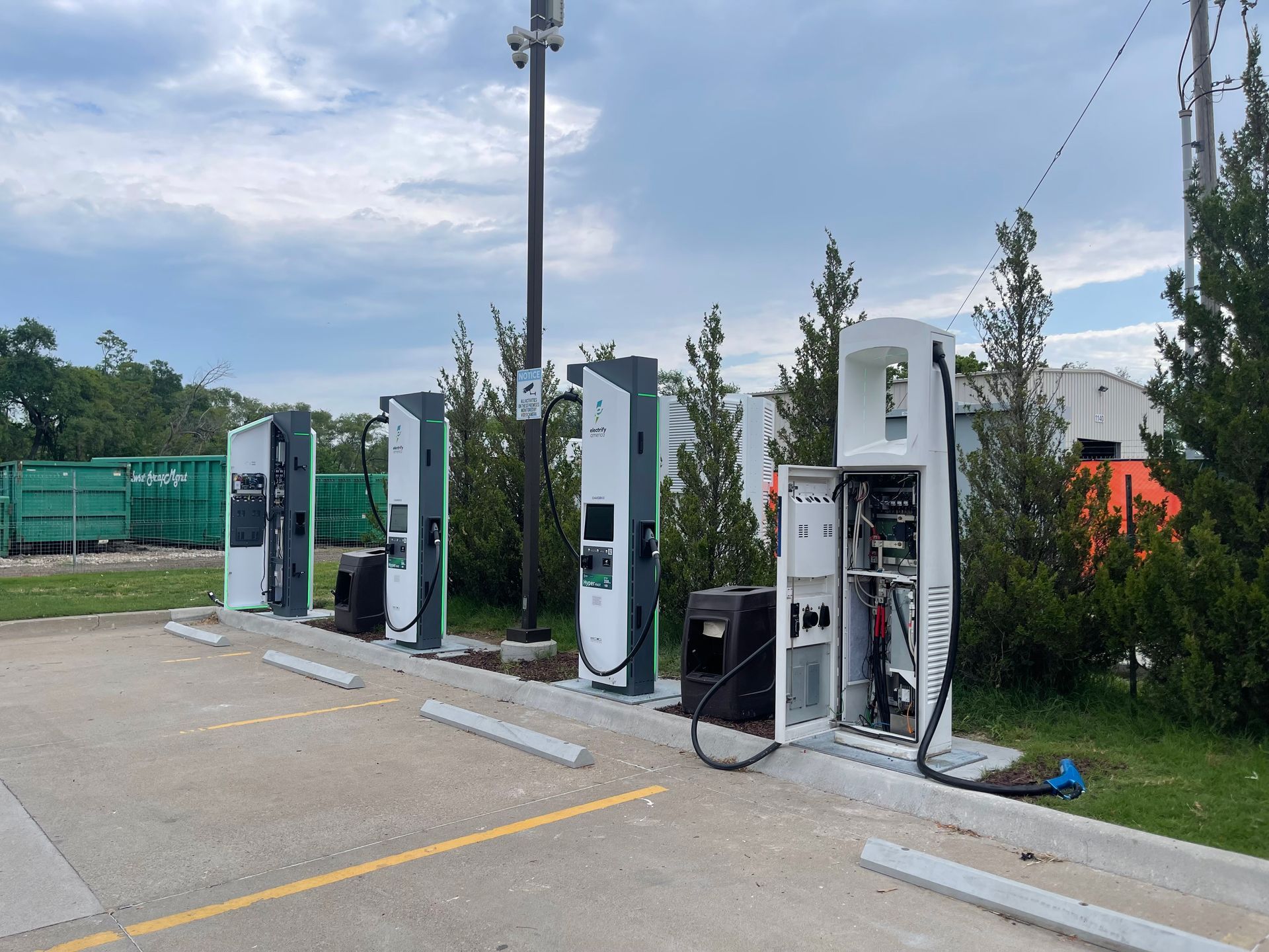 Row of electric vehicle charging stations beside a parking lot, with trees and a cloudy sky.