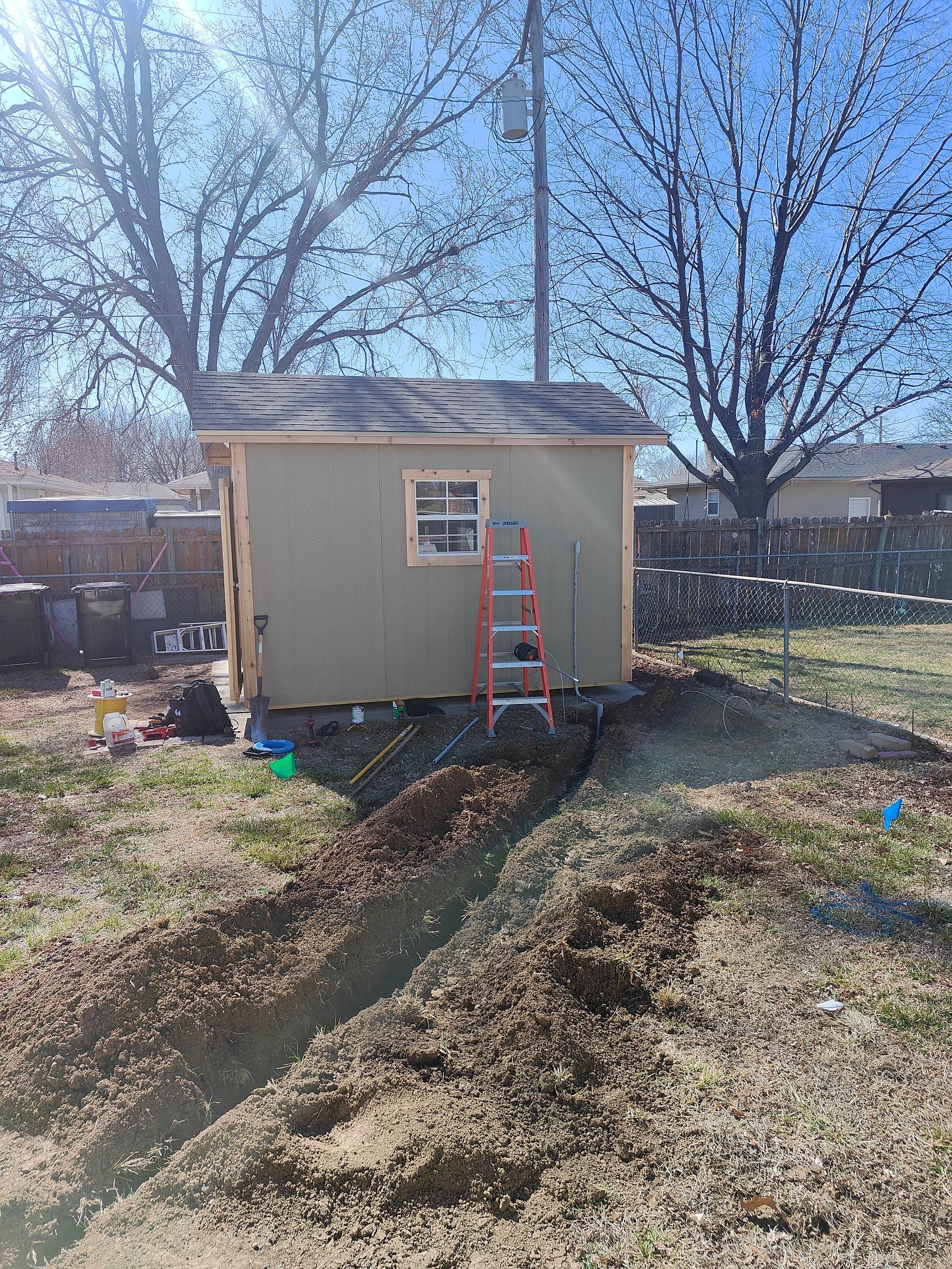 Small beige shed in a yard with a red ladder leaning against it and a mound of dirt in front.
