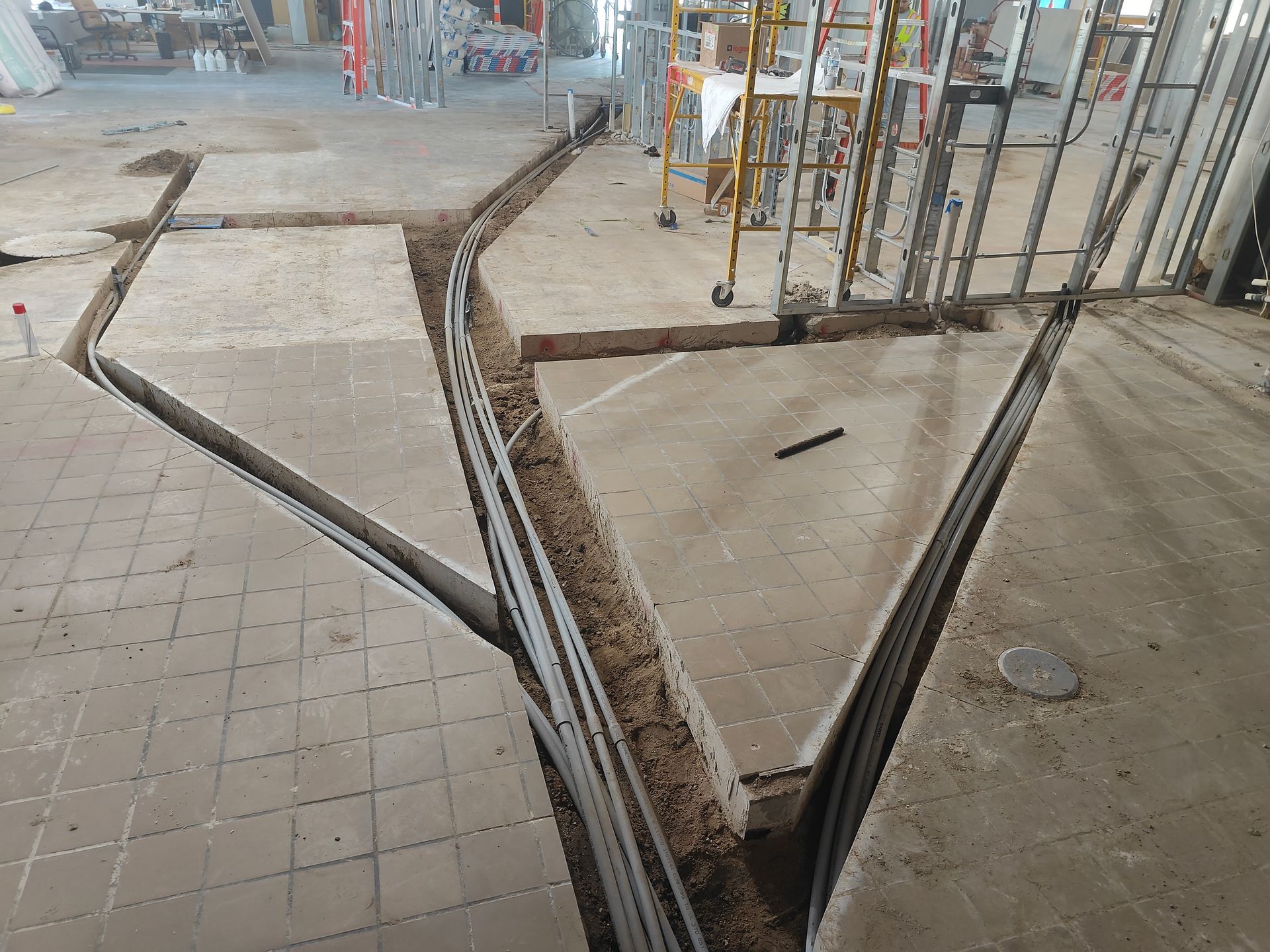 Damaged sidewalk with open concrete trenches and exposed cables near a construction area