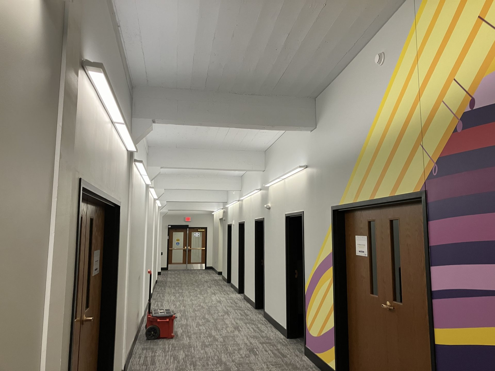 Bright school hallway with white walls, wooden doors, and colorful mural accents, with a small red cart on the floor.