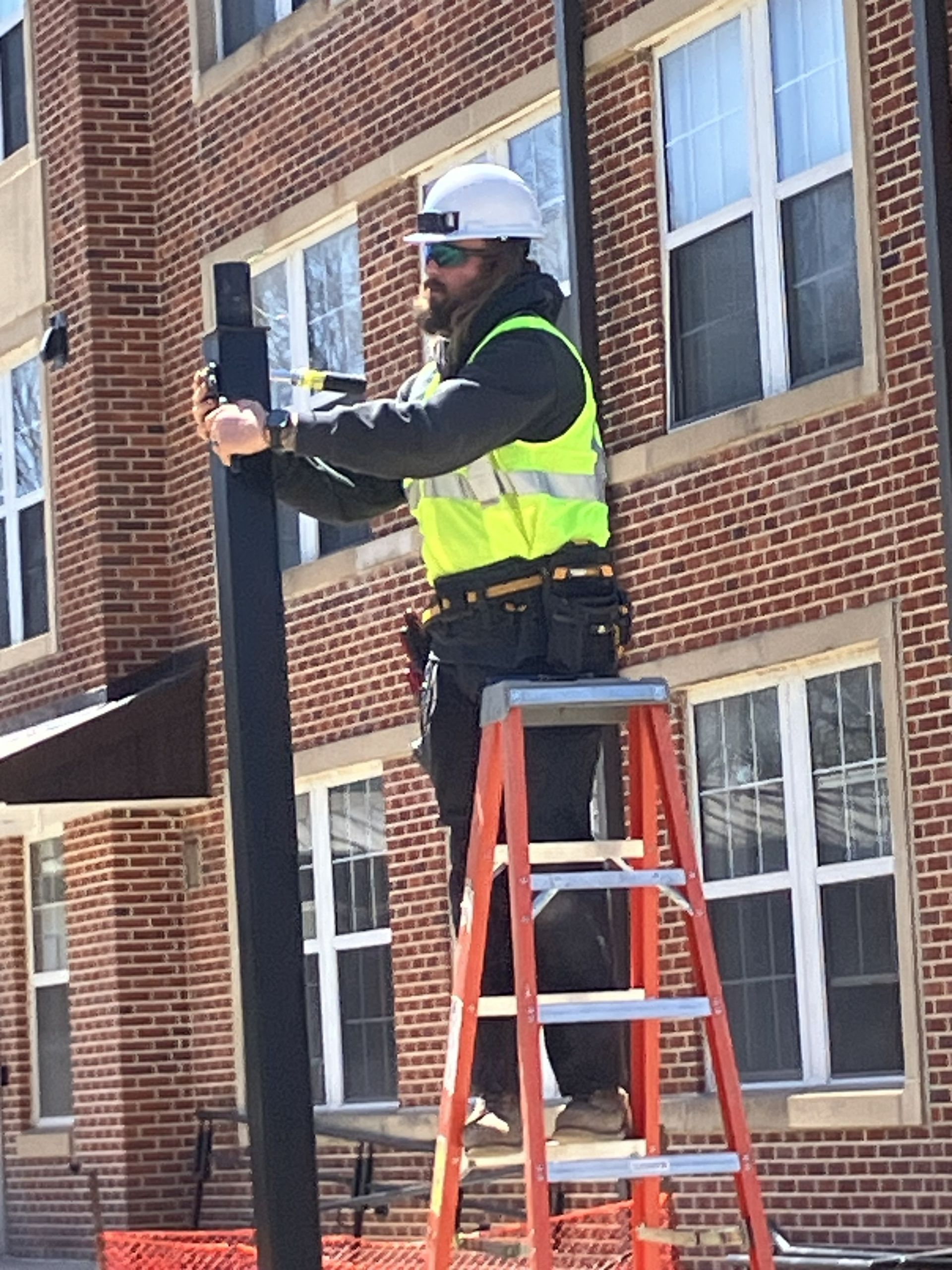 Worker in hard hat and safety vest on a ladder measuring a black pole beside a brick building