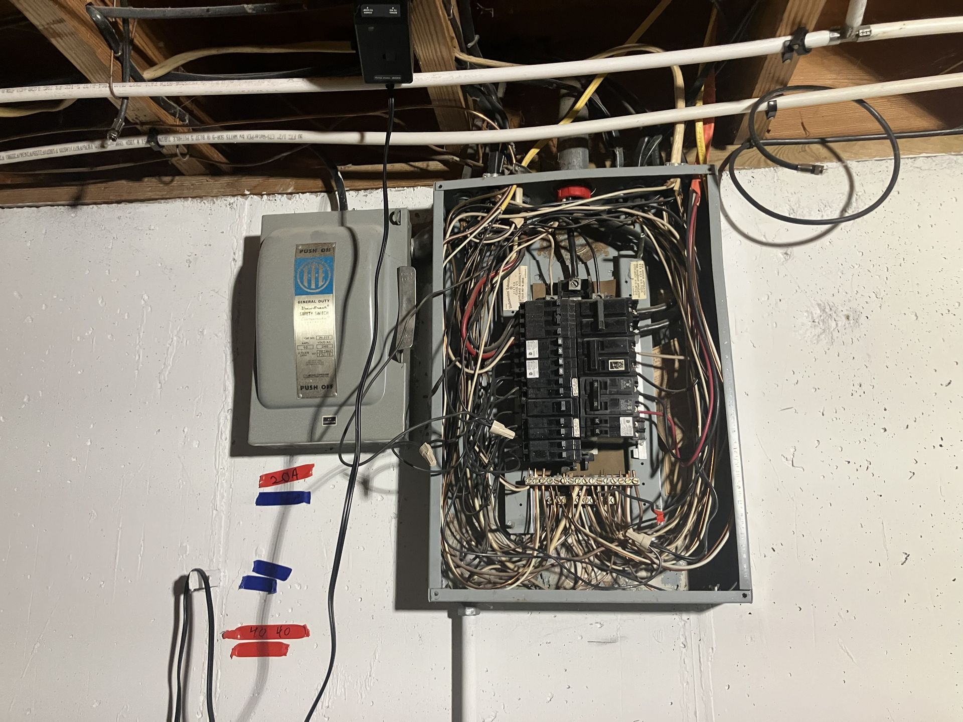 Basement wall with electrical panels, exposed wiring, and labeled circuit breakers.