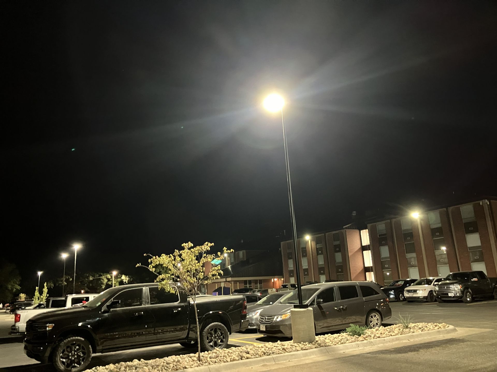 Night parking lot with cars, a bright streetlamp, and a brick building in the background.
