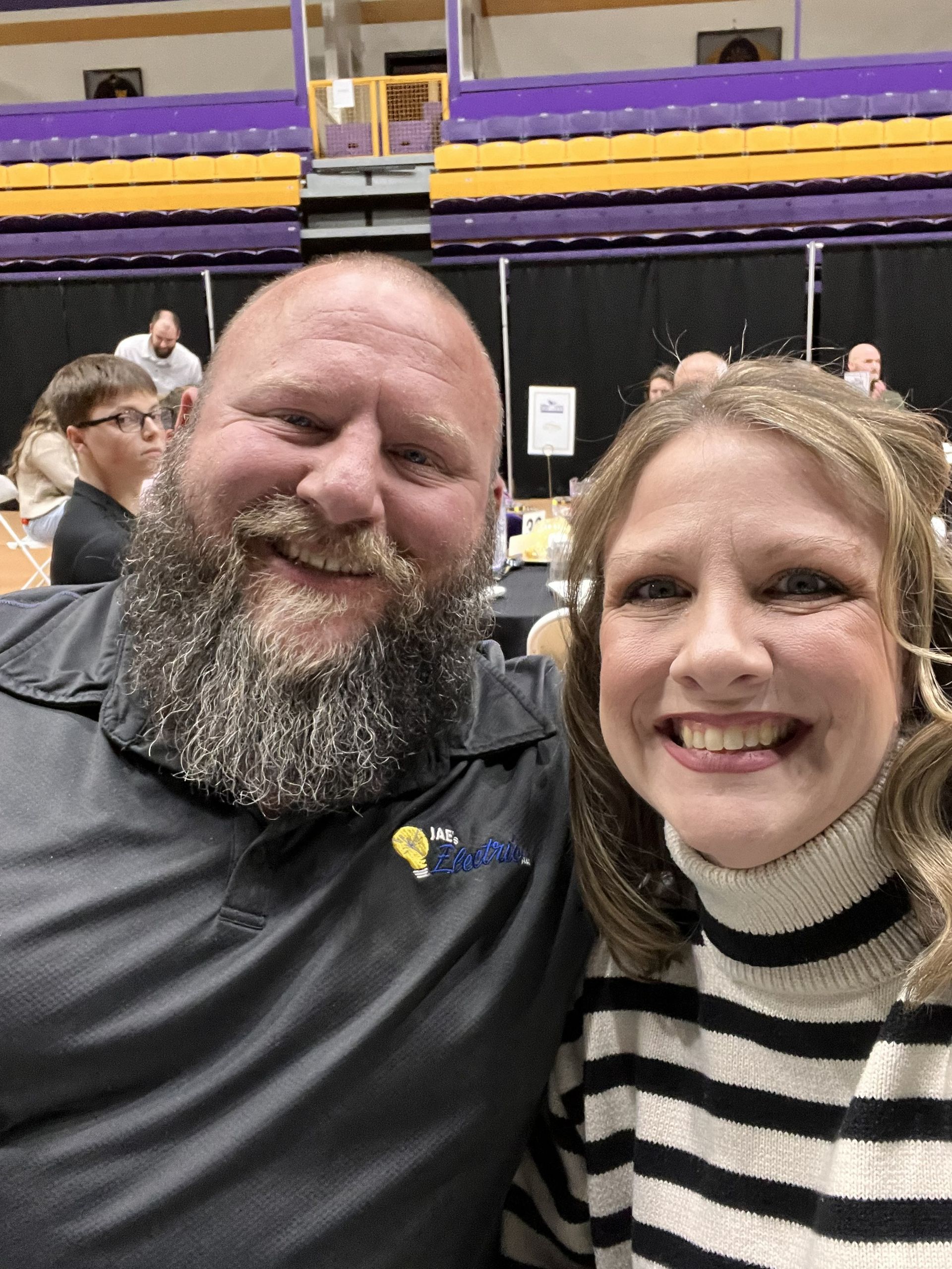 Two smiling people posing indoors in a stadium, with purple seats in the background.