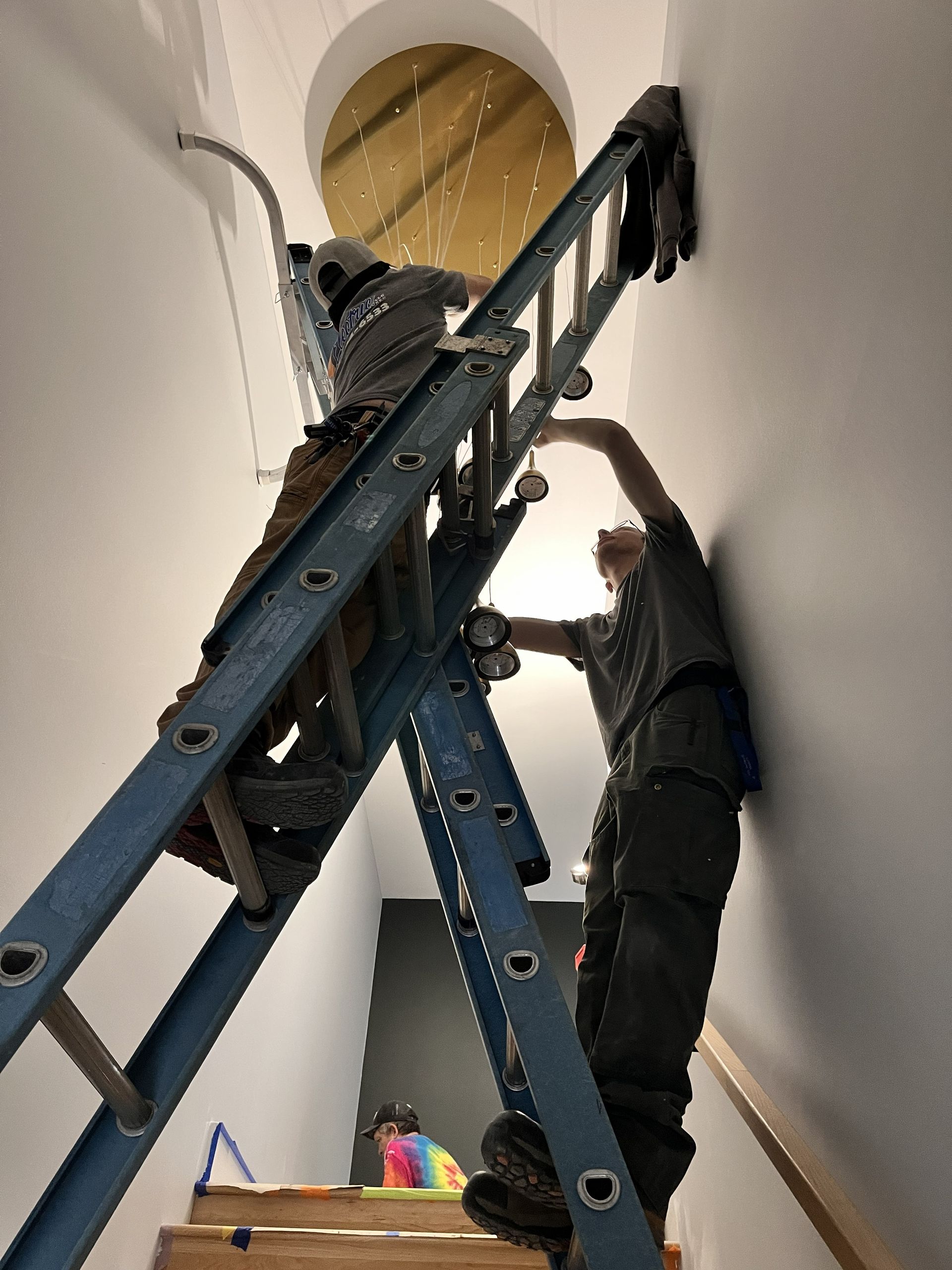 Two workers on ladders installing equipment in a tall stairwell with a round skylight