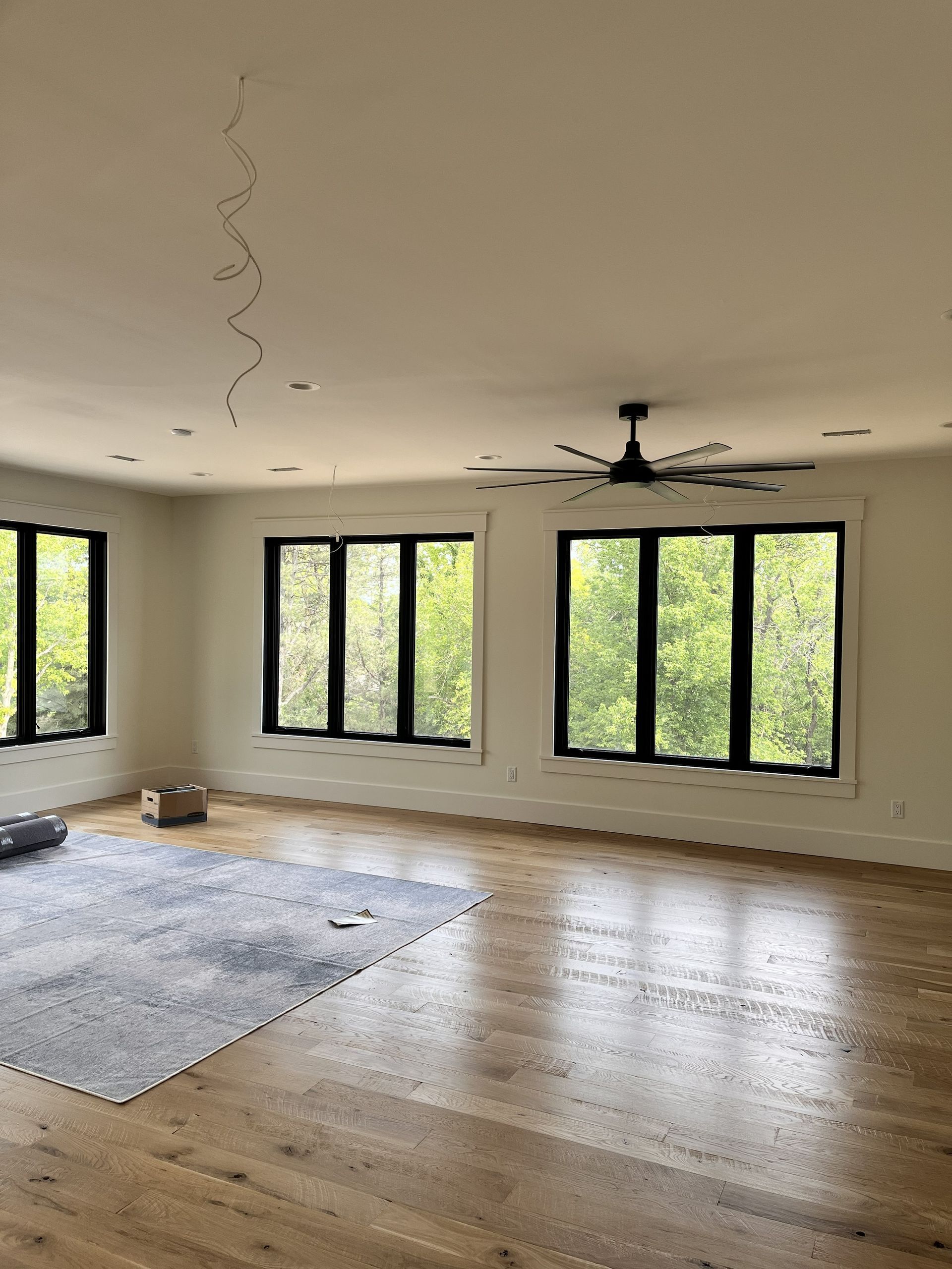 Empty modern living room with hardwood floors, large black-framed windows, and a ceiling fan under construction
