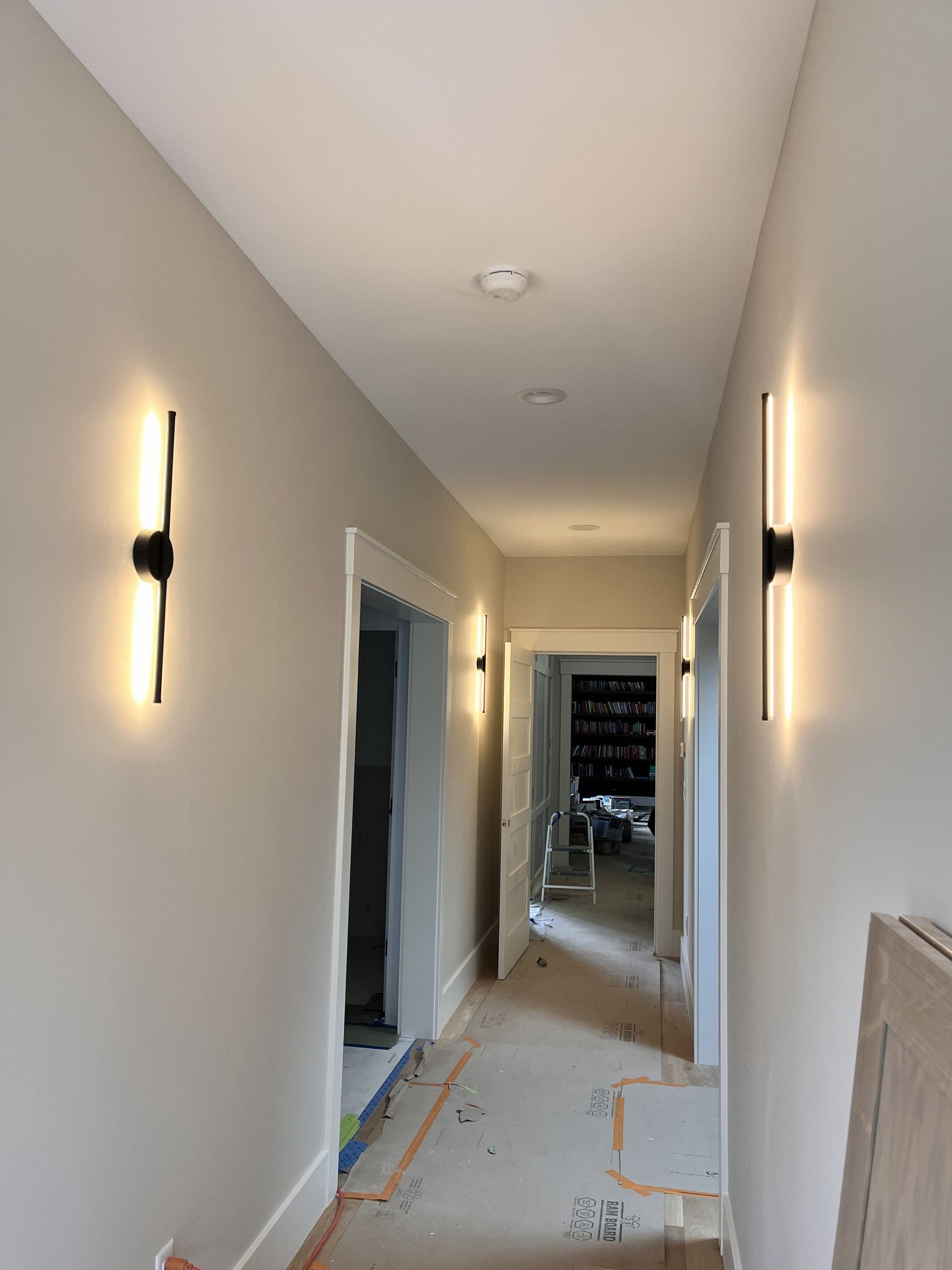 Narrow hallway under construction with wall sconces, unfinished floor, and doorway at the far end.