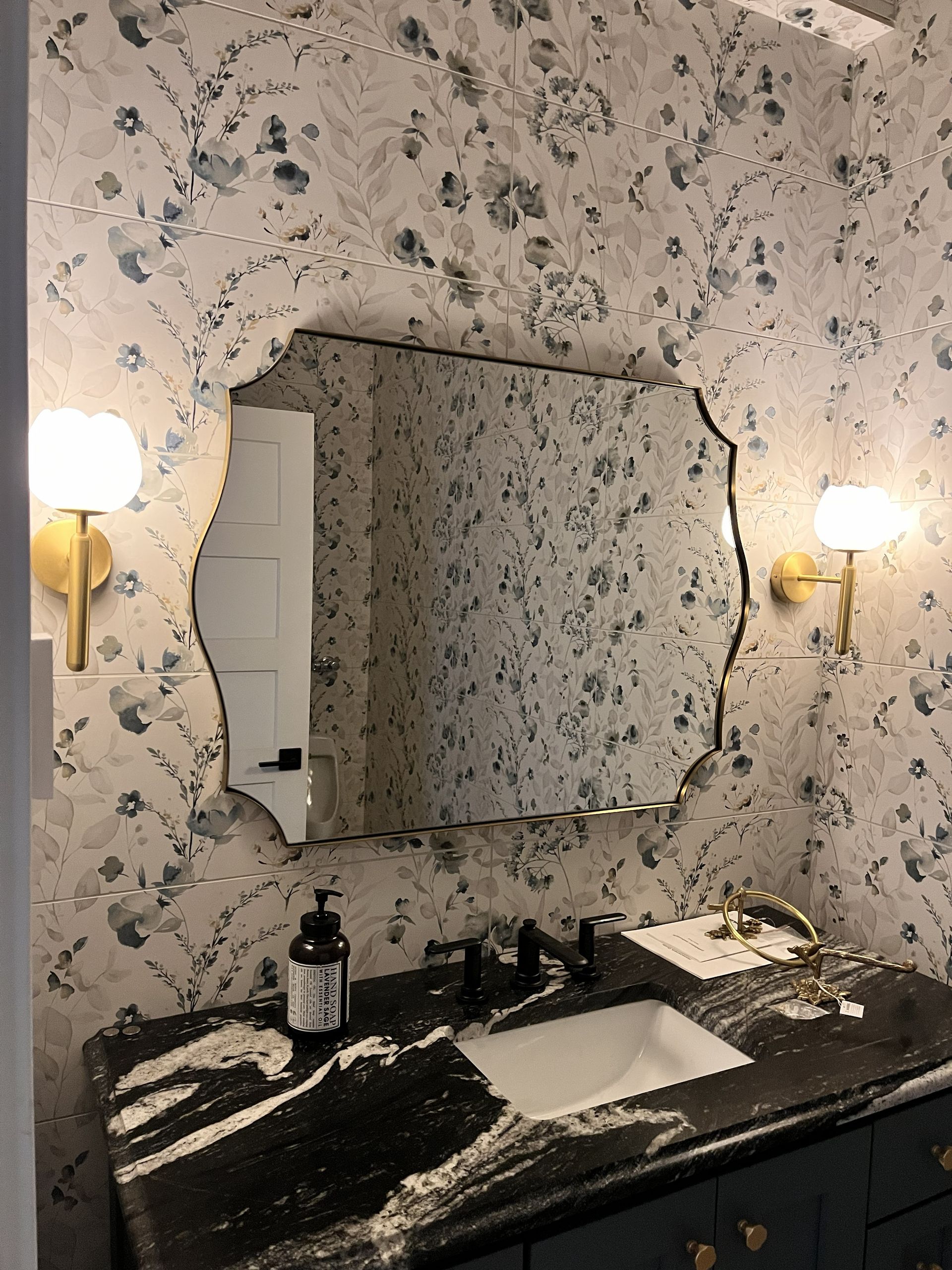 Floral wallpaper bathroom vanity with lit sconces, mirror, and black countertop around a white sink