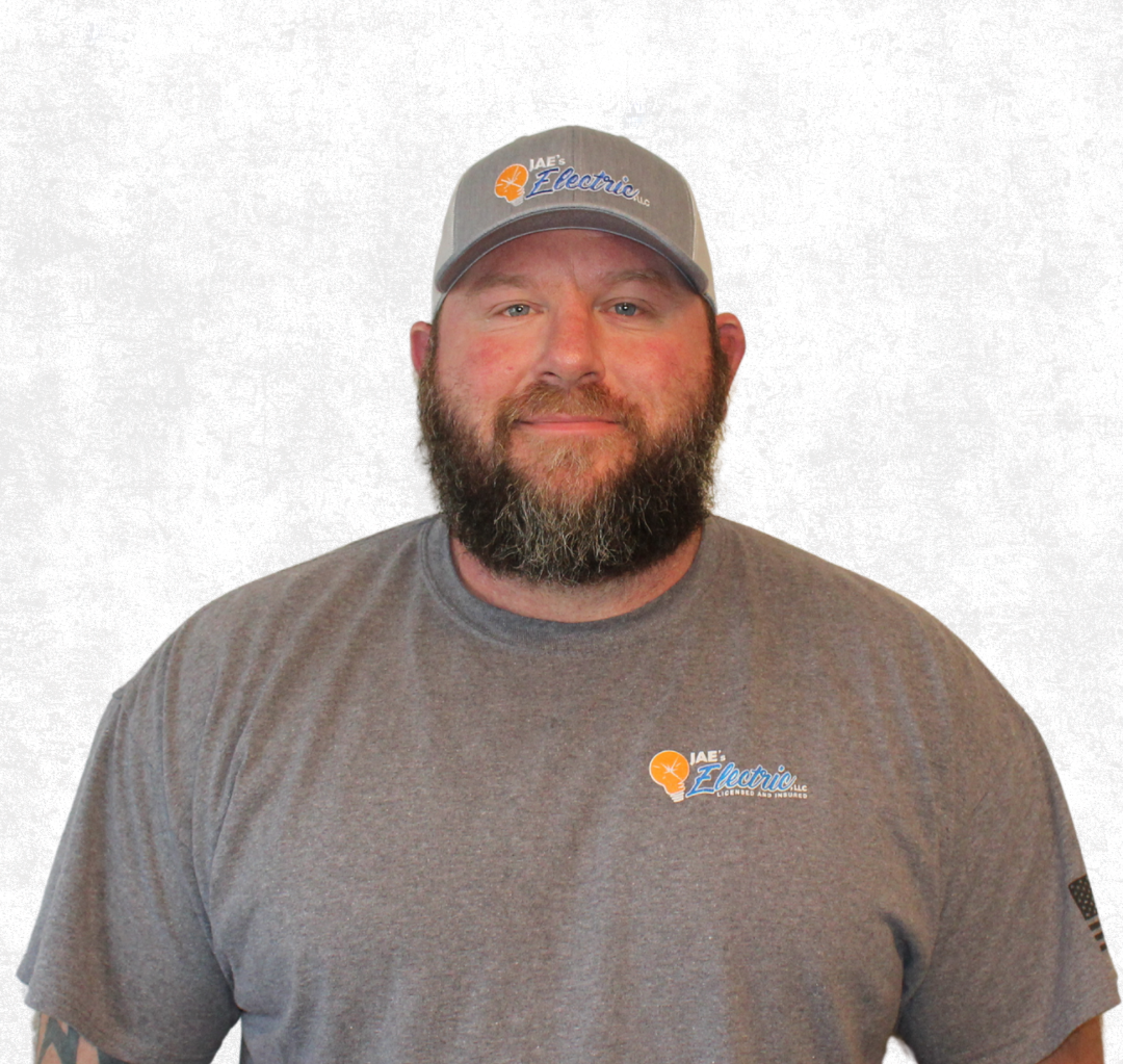 Man in gray shirt and cap with beard, standing against a plain light background.
