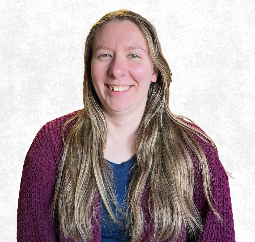 Smiling person with long blonde hair wearing a purple cardigan against a light background