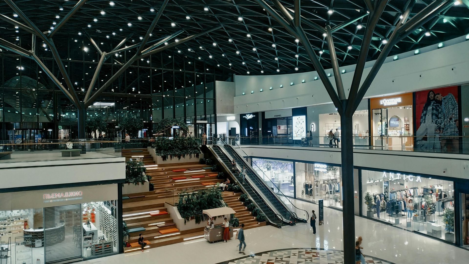 Modern indoor mall atrium with escalators, glass railings, and shoppers under a high steel-truss ceiling