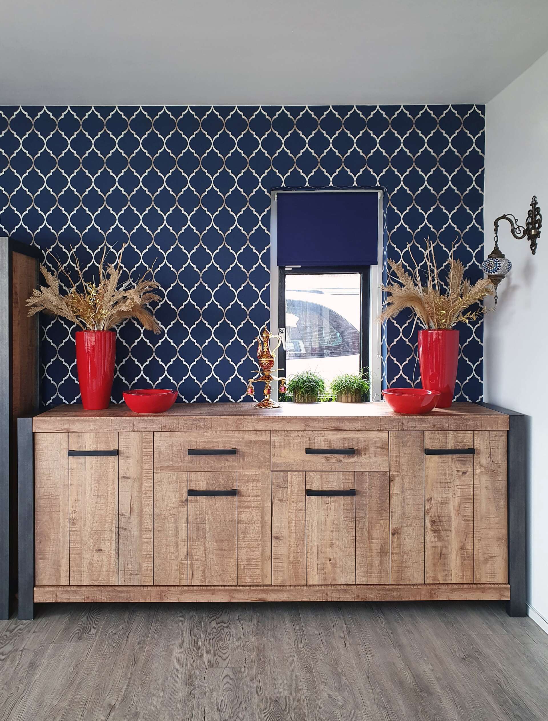Een rustiek houten dressoir met zwart beslag staat voor een blauw patroonmuur met een raam, geflankeerd door rode vazen.