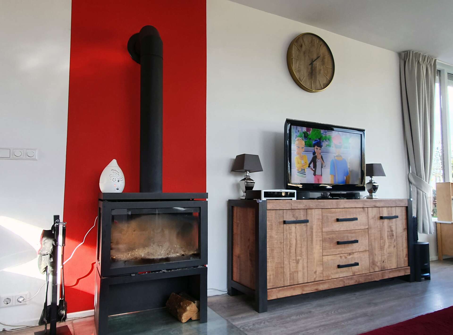 Een woonkamer met een zwarte houtkachel tegen een felrode muur, naast een rustiek houten tv-meubel met een televisie.