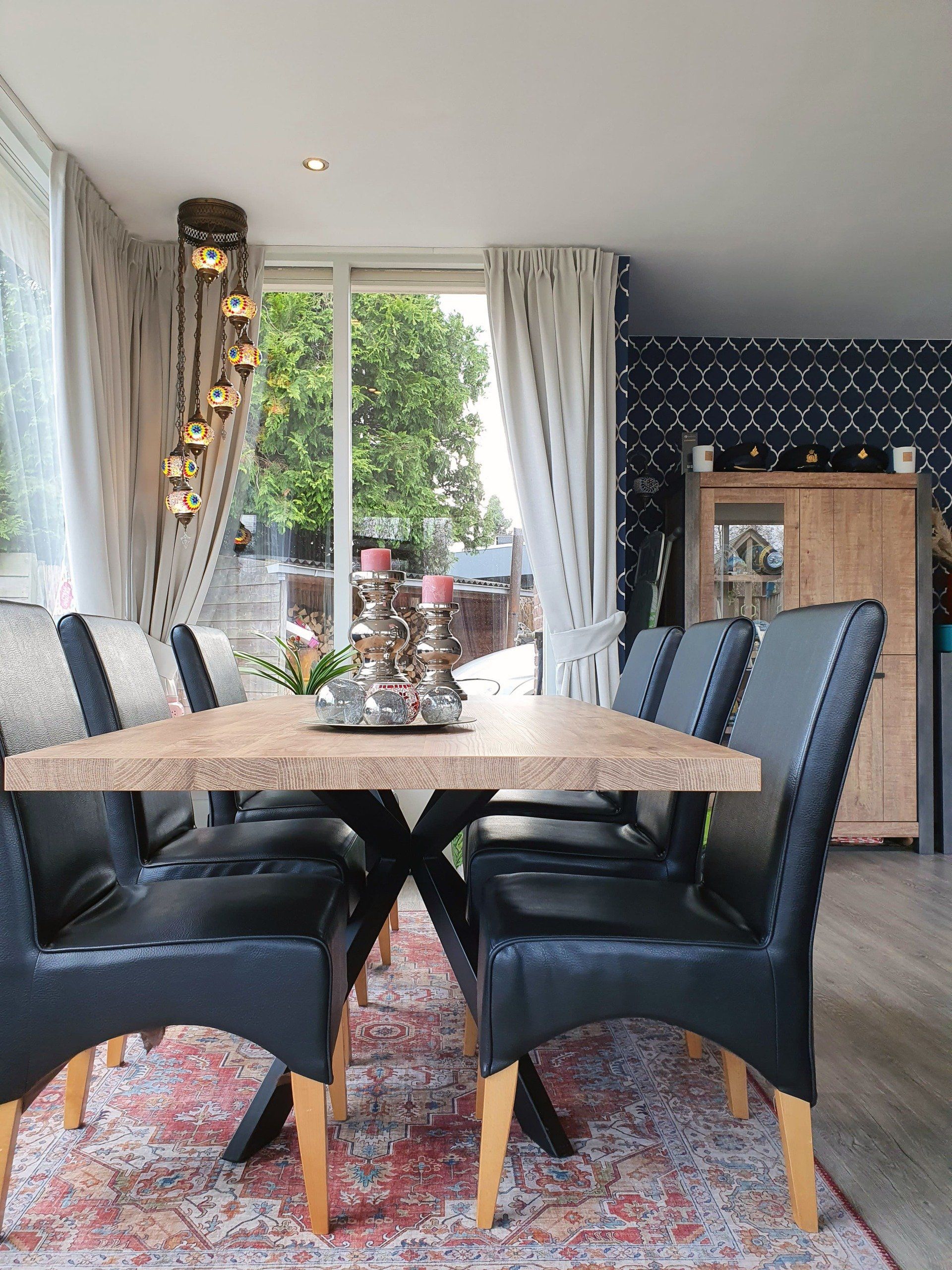 Een eetkamer met een lange houten tafel, zwarte gestoffeerde stoelen en een hanglamp bij een licht raam.