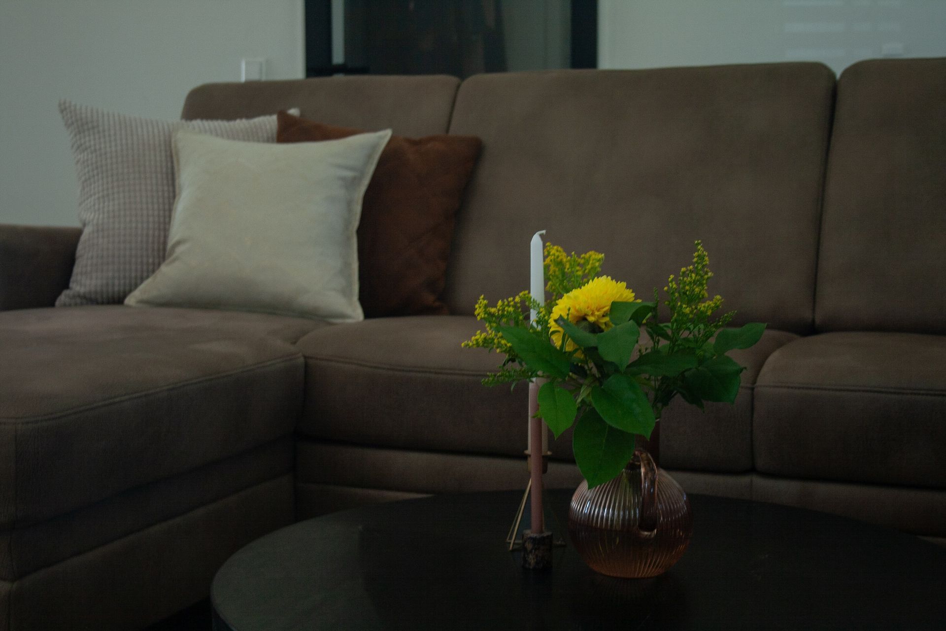 Een close-up van een klein glazen vaasje met gele bloemen en een dun kaarsje op een donkere ronde tafel voor een bruine bank.