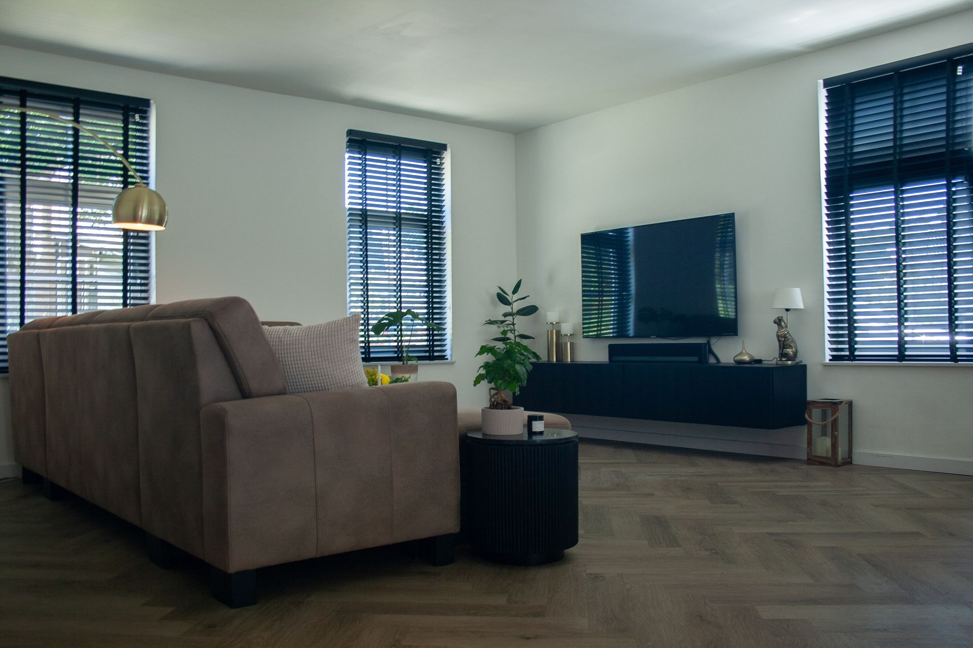 Moderne woonkamer met een beige bank en donkere raamjaloezieën.