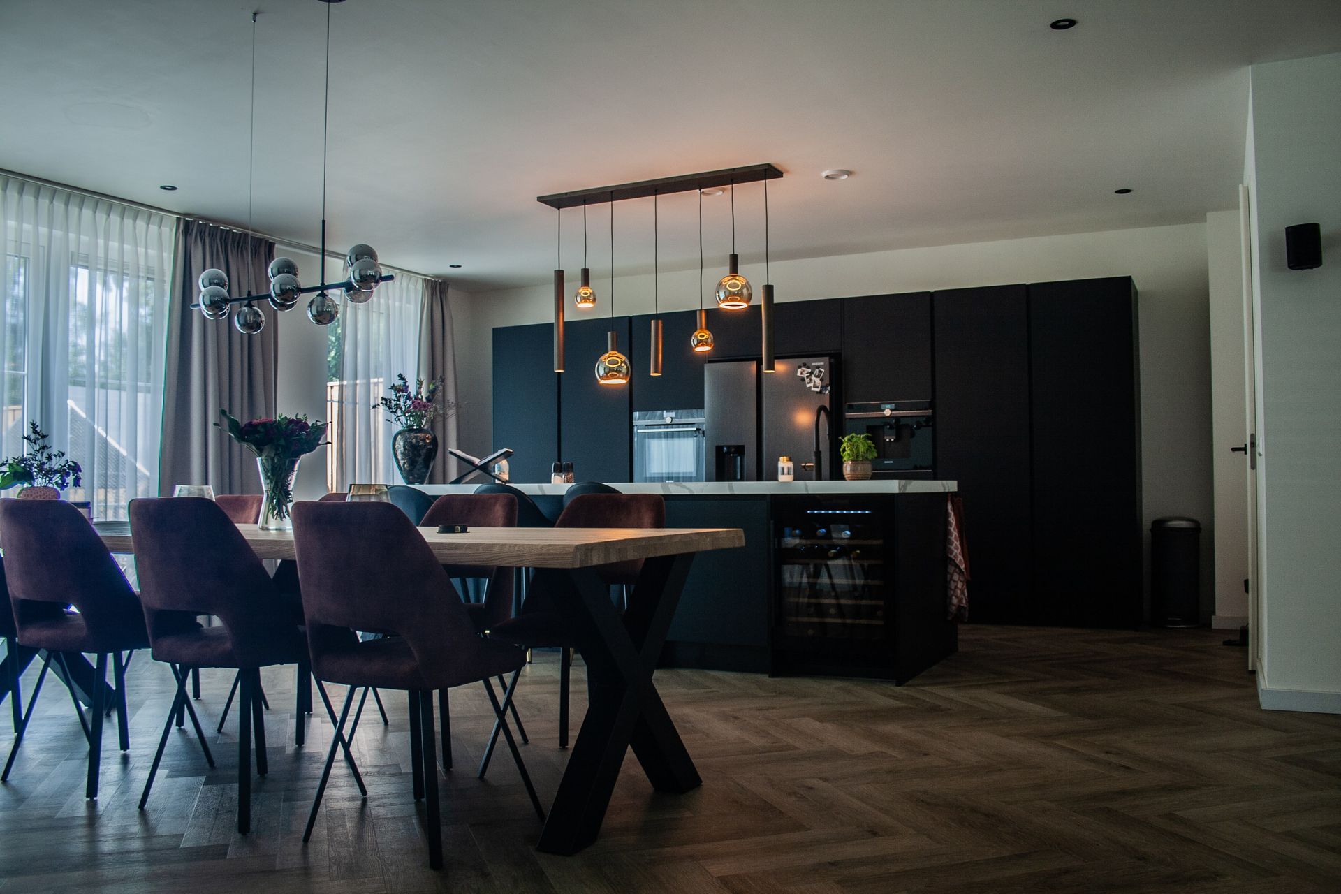 Een moderne eet- en keukenruimte met een houten tafel, donkere fluwelen stoelen, zwarte kasten en industriële hanglampen.
