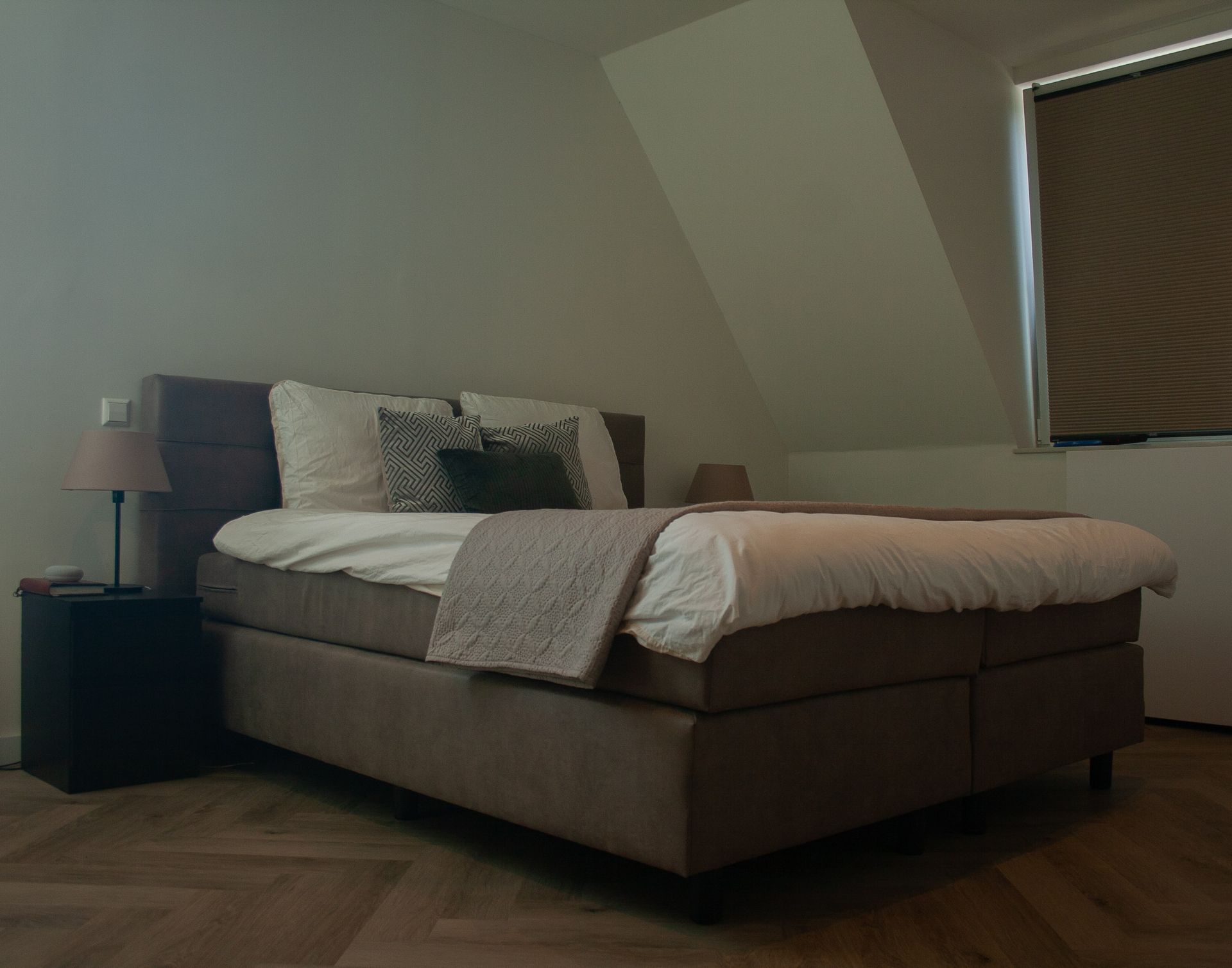 Een slaapkamer met een gestoffeerd bed met licht beddengoed, een nachtkastje, een lamp en een raam met jaloezieën.