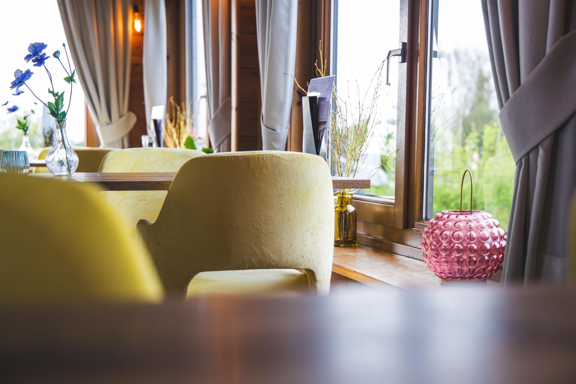 Een wazige voorgrond leidt naar gele fauteuils in een gezellig restaurant met gordijnen en een roze lamp bij het raam.
