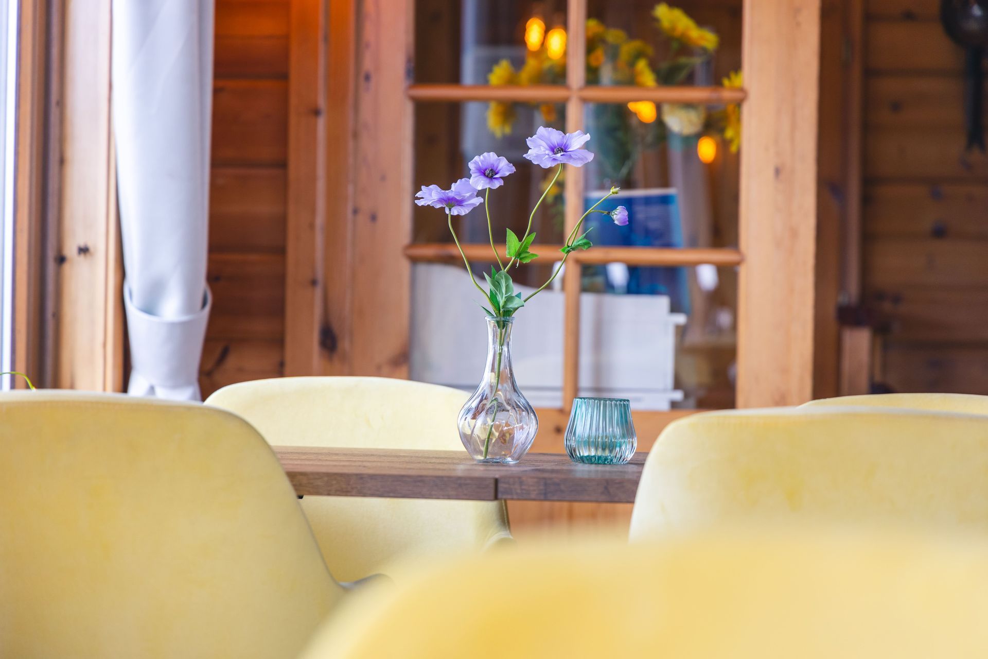 In een rustieke, zonnige kamer staat een vaas met paarse bloemen en een klein glas op een houten tafel tussen gele stoelen.