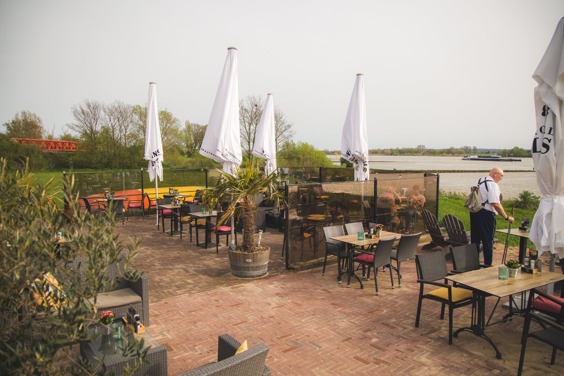 Buitenterras van een restaurant aan de rivier met lege tafels, stoelen en grote witte parasols op een grindondergrond.