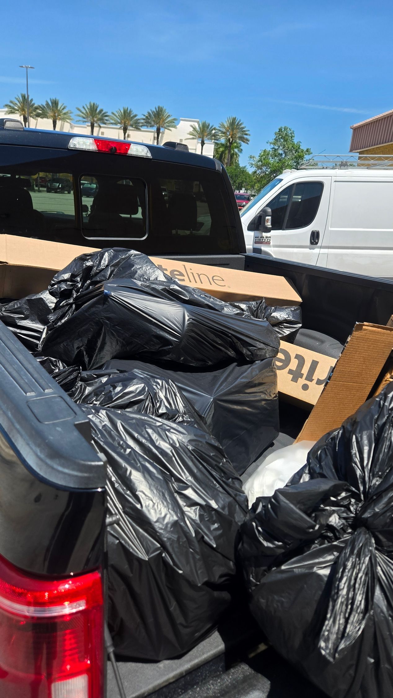 Black trash bags piled in the back of a pickup truck in a parking lot with a white van nearby