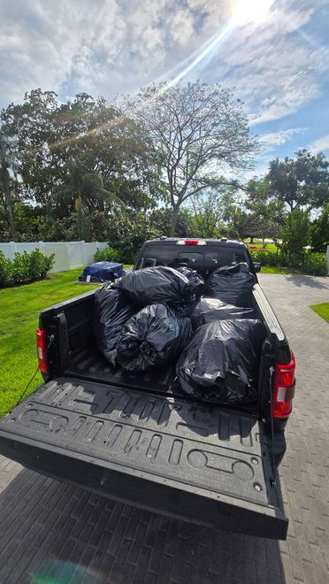 Pickup truck bed full of black trash bags parked in a sunny driveway under a blue sky.