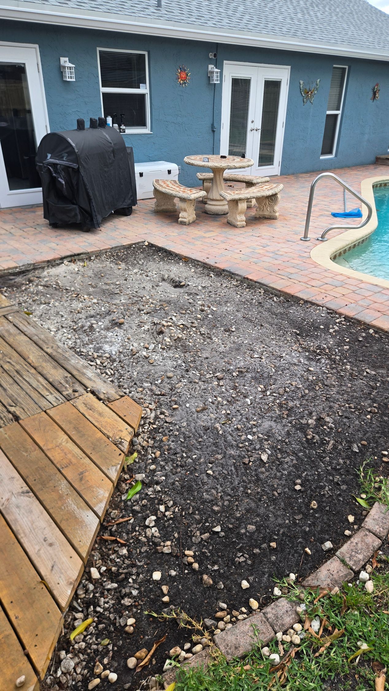 Backyard patio with blue house, grill, table, pool, and a gravel area beside wooden decking.