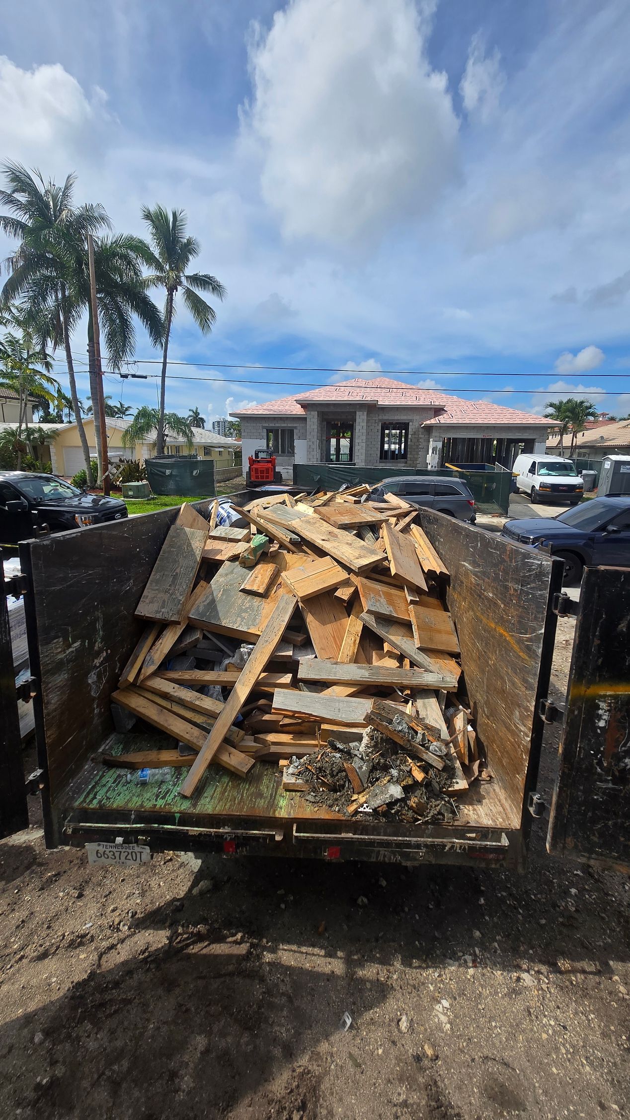 Trailer loaded with broken wooden debris on a muddy lot, with a house and palm trees in the background