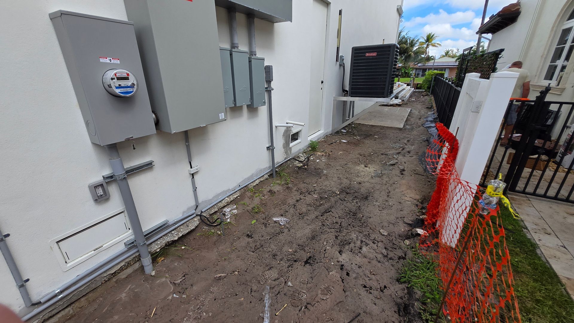 Narrow dirt alley beside gray utility boxes and a white building, bordered by orange safety fencing