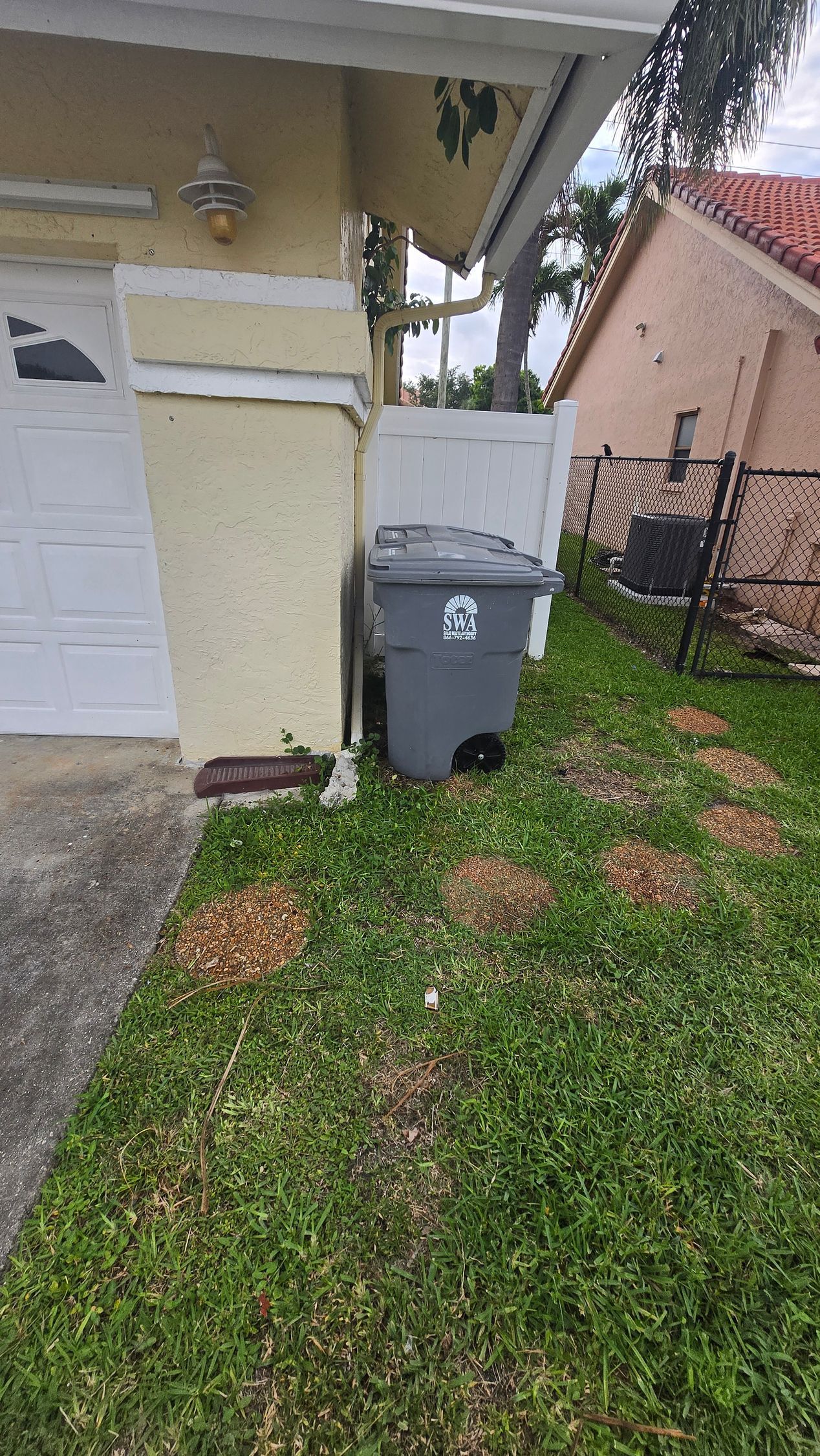 Gray trash bin beside a cream-colored house and garage on a grassy side yard.