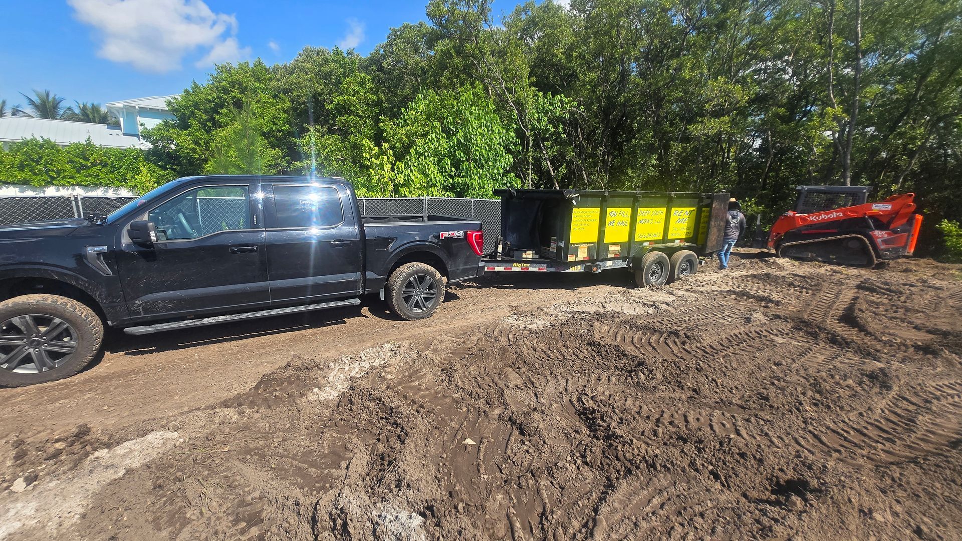 Black pickup truck with trailer and yellow bins on muddy ground near trees, with red excavator in the background