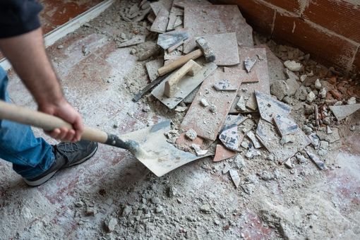 Person shoveling broken tiles and rubble in a dusty corner of a room