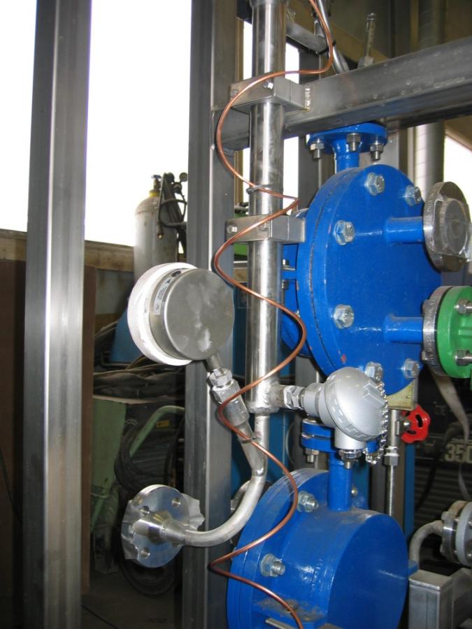Industriutstyr med blå tanker, kobberrør og diverse målere og ventiler i en metallramme.
