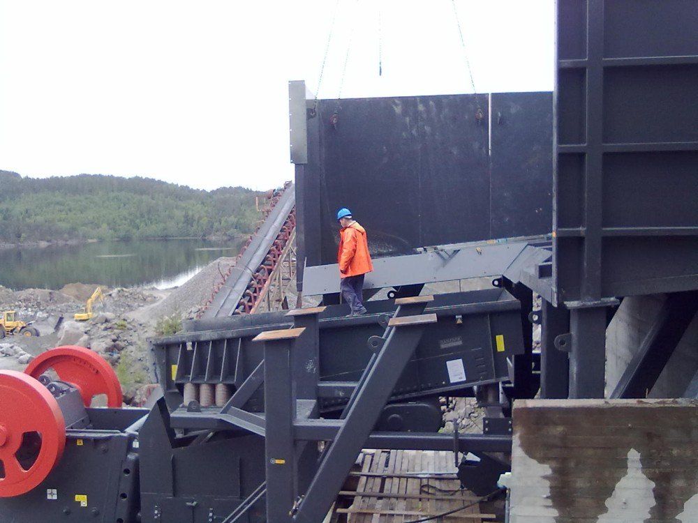 Mann i oransje jakke inspiserer en industriell steinknuser, med et transportbånd og naturlige omgivelser synlige.
