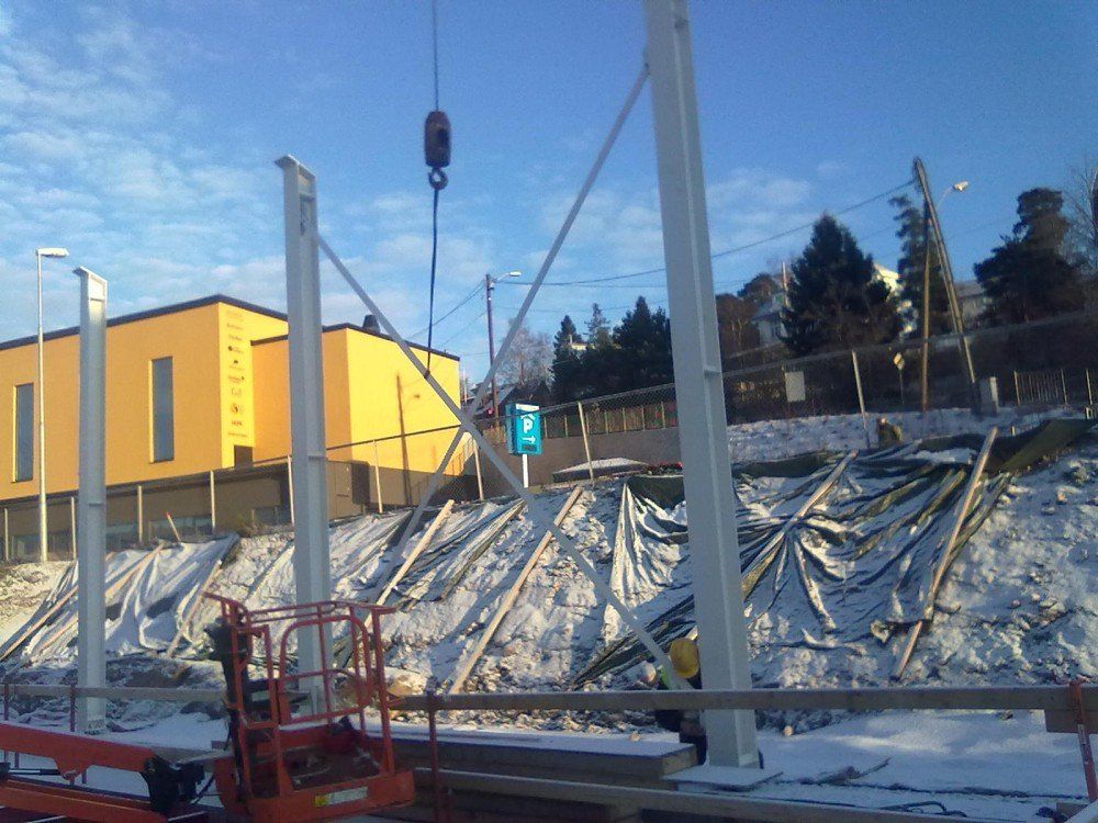 Byggeplass med kran som løfter stålbjelker, mot en gul bygning og en snødekt åsside.