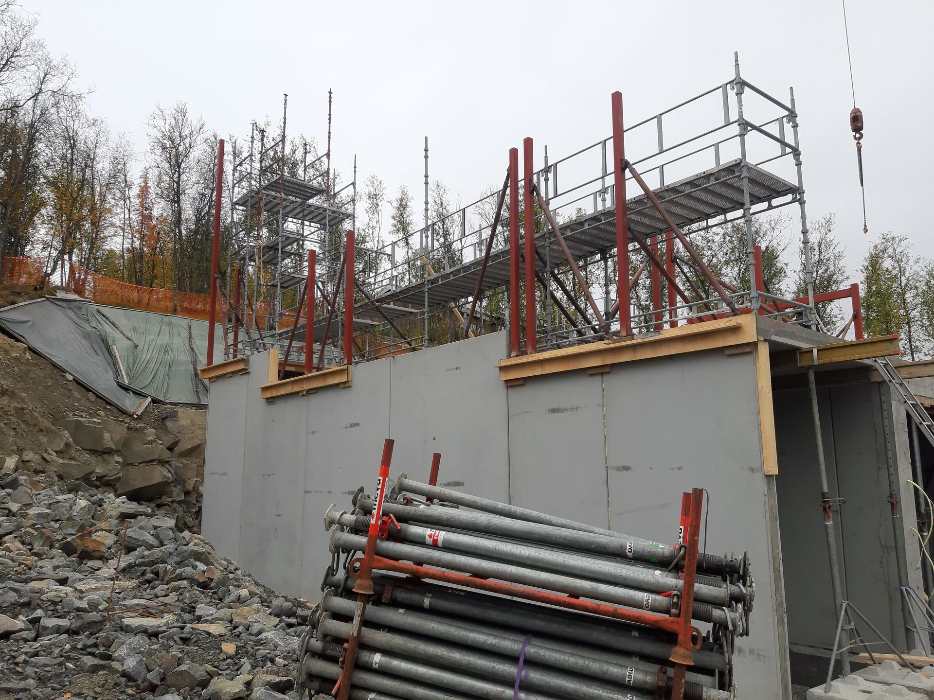 Byggeplass med grå betongvegg, stillas og stablede metallstolper.