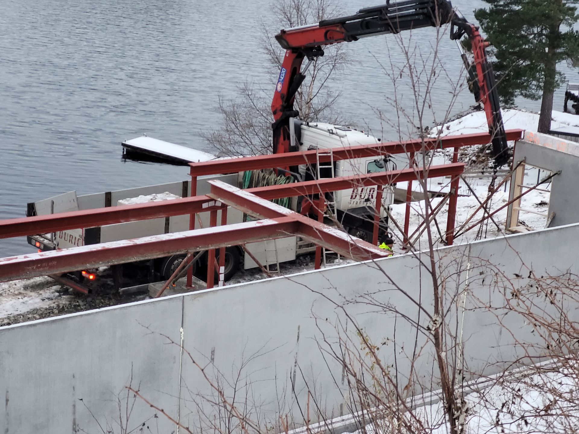 Kran losser metallbjelker ved siden av en snødekt innsjø. Hvit lastebil parkert nær en betongvegg.
