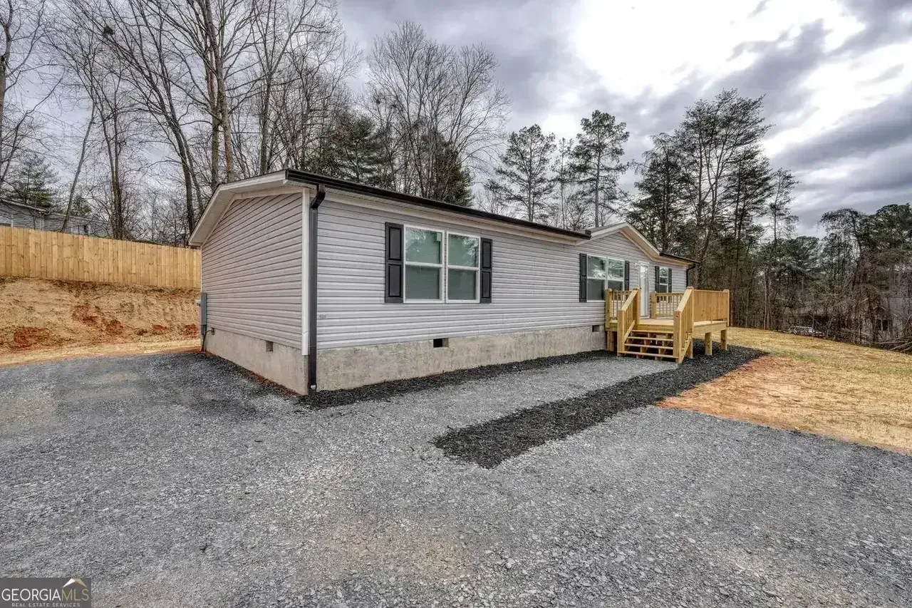 Small gray manufactured home on gravel lot with wood fence, trees, and cloudy sky