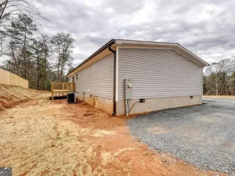 Single-story beige house with gravel driveway and wooded yard under cloudy sky