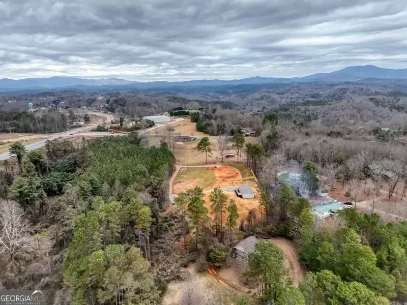 Aerial view of a rural landscape with dirt roads, green trees, cleared land, and distant mountains under cloudy skies