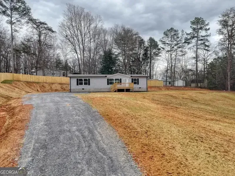 Small gray house with a wooden porch at the end of a gravel driveway on a wooded lot under cloudy sky