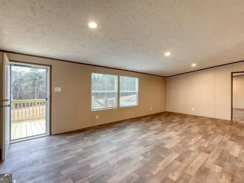 Empty beige living room with wood-look flooring, recessed lights, and a sliding glass door to a deck.