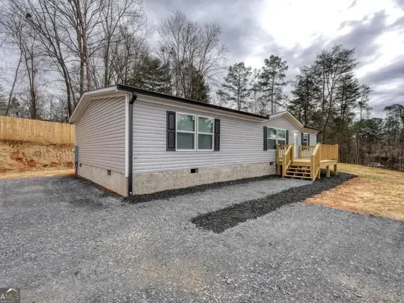 Single-wide gray mobile home with a small wooden deck on a gravel lot, surrounded by trees