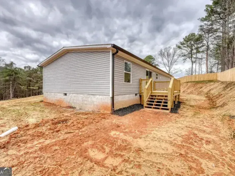 Single-wide mobile home with gray siding and wooden steps on a bare dirt lot under cloudy skies