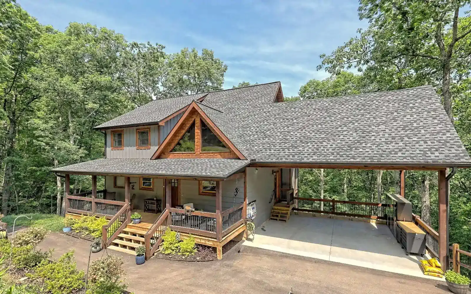 Rustic two-story cabin with wraparound porch and open carport, surrounded by trees and landscaping.